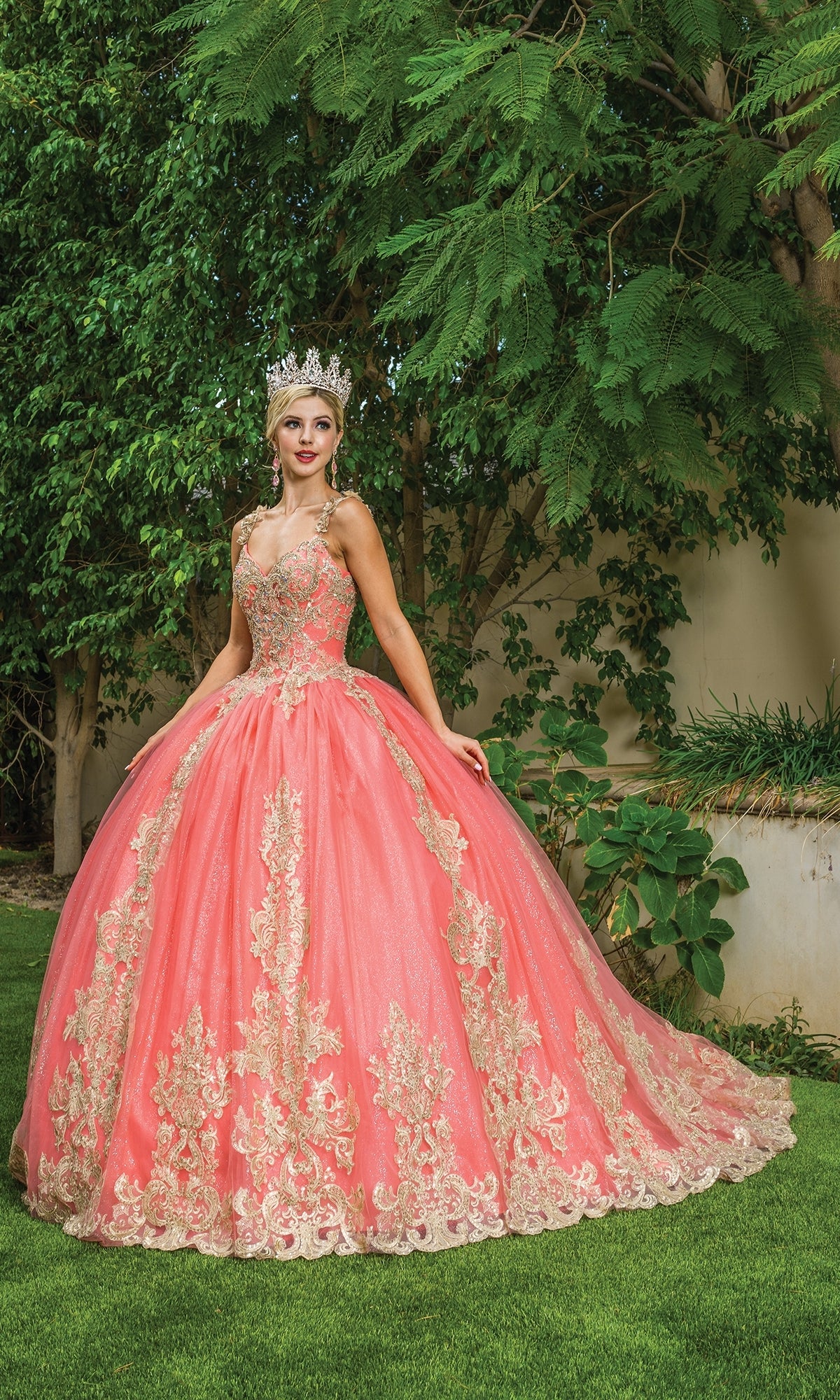 A young woman wearing a crown and the Coral Quinceanera Dress 1629 with gold lace embroidery stands on grass in front of green trees and bushes.